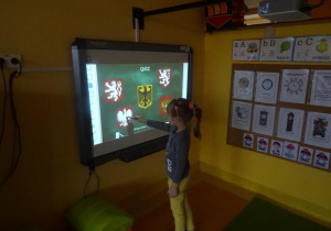 Dziewczynka stoi przy tablicy interaktywnej, w ręku trzyma wskaźnik skierowany na godło Polski.